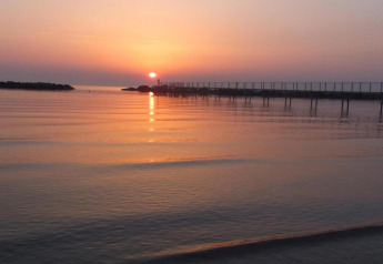 Atardecer sobre aguas tranquilas visto desde una casa móvil, con un largo muelle y cielo pastel.