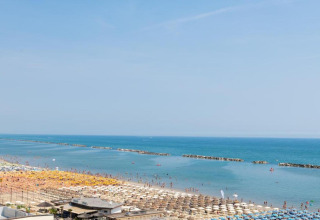 Uitzicht vanuit een stacaravan op een druk strand met kleurrijke parasols en een rustige blauwe zee.