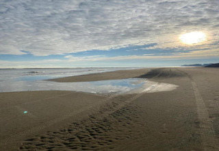 Foto van een rustige zandstrand met bandensporen, een waterplas, zon en dramatische bewolking.