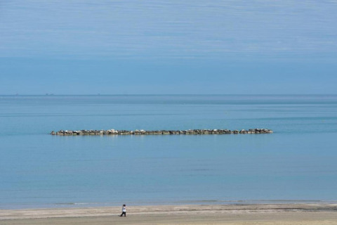 Een persoon loopt op het strand bij een stacaravan, met een kalme zee en een stenen golfbreker op de achtergrond.