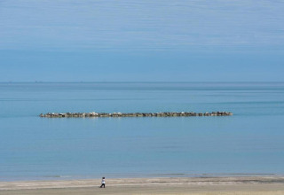 Eine Person spaziert am Strand nahe einem Mobilheim, mit ruhigem Wasser und einer Steinmole im Hintergrund.