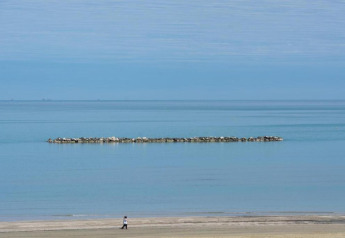 En person går på stranden foran et mobilhome, med roligt blåt hav og en stenmole i baggrunden.