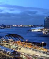 Vista de una ciudad moderna junto al río al anochecer, con estación de tren y edificios iluminados.