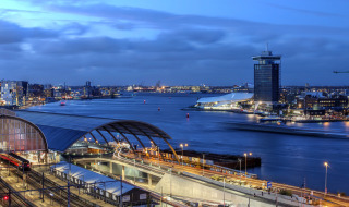 Vista de una ciudad moderna junto al río al anochecer, con estación de tren y edificios iluminados.