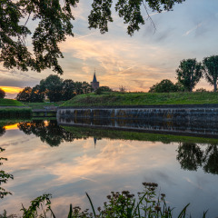 Foto de los alrededores de Naarden, en Holanda Septentrional, Países Bajos, con canal, árboles y atardecer reflejado.