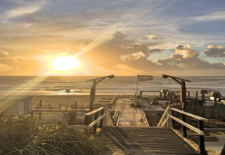 Tramonto sul mare visto dal pontile di un lodge, con sabbia, erba e alcune strutture sulla spiaggia.
