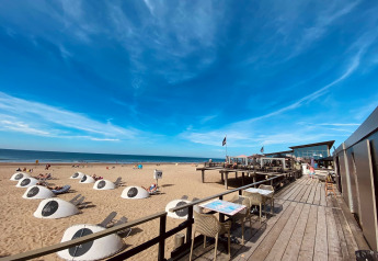 Sunny beach with modern lodge, outdoor café seating, unique white cabanas, and clear blue sky by the sea.