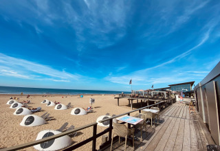 Spiaggia soleggiata con lodge moderno, tavoli all’aperto, cupole bianche e cielo azzurro vicino al mare.