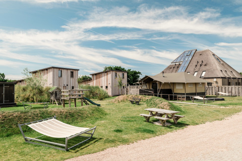 Modern lodgecomplex met houten lodges, picknicktafels, speelzone en hangmat onder een blauwe hemel.