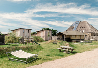 Modern lodgecomplex met houten lodges, picknicktafels, speelzone en hangmat onder een blauwe hemel.