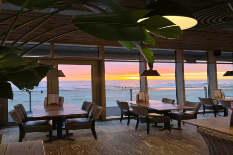 Interior view of a beachfront restaurant with empty tables, large windows, and a sunset over the ocean outside.