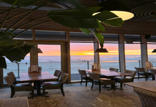 Binnenzicht van een strandlodge met lege tafels, grote ramen en een prachtige zonsondergang aan zee.