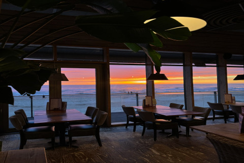 Vista interna di un ristorante sul mare con grandi finestre, tavoli vuoti e un tramonto sull’oceano.
