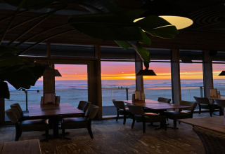 Vista interna di un ristorante sul mare con grandi finestre, tavoli vuoti e un tramonto sull’oceano.