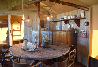 Rustic dining area in Kidz Lodge, Netherlands with wooden round table, chairs and cozy kitchen shelves.