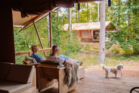 Two people relax on the porch of a Veluwe Lodge safari tent with a small dog and forest scenery around.