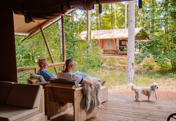 Dos personas se relajan en la terraza de una tienda safari Veluwe Lodge con un pequeño perro y naturaleza.