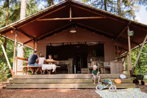 Gezin ontspant op de veranda van de Veluwe Lodge safaritent op Camping Samoza in het Nederlandse bos.