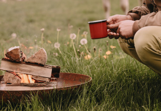 Personne accroupie sur l'herbe avec une tasse rouge près d'un feu de camp entouré de fleurs sauvages.