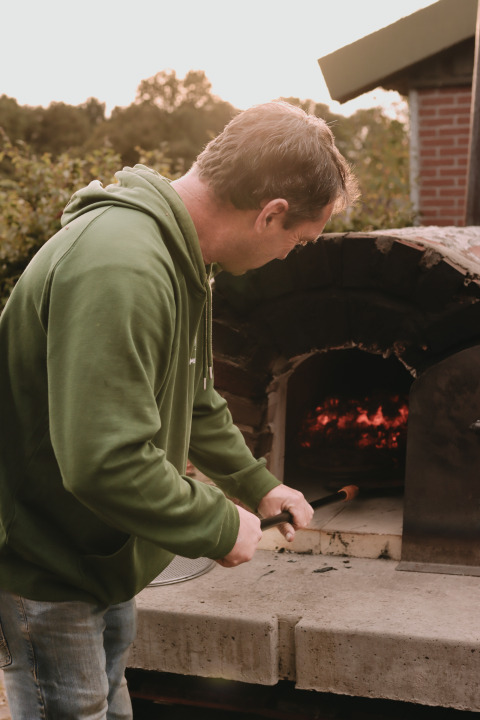 Man in green hoodie cooking by wood-fired oven at Feather Down De Oude Melkstal, North-Brabant, Netherlands.