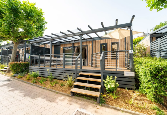Moderne lodge bij Next L EVO met houten gevel, overdekt terras en tuintje in het zonlicht.