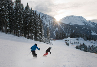 To personer står på ski ned ad en piste ved Almhütte Nase, omgivet af sneklædte bjerge og skove i Østrig.