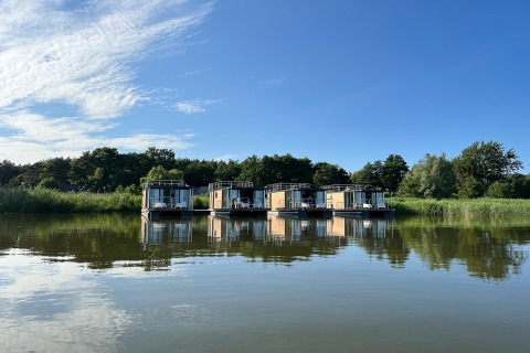 Vier moderne woonboten voor twee personen aan de oever, omgeven door groen en weerspiegeld in het water.