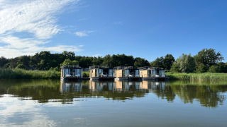 Vier moderne Hausboote für zwei Personen liegen am Ufer, umgeben von Bäumen, Himmel und Wasser.