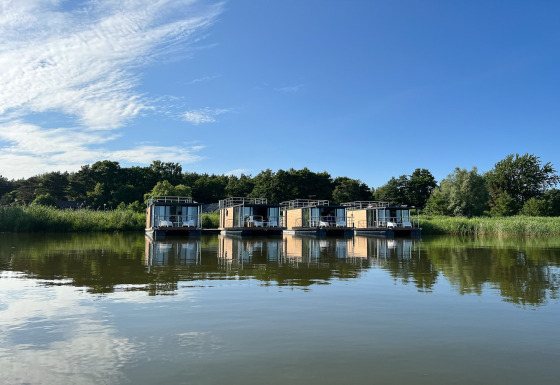Vier moderne woonboten voor twee personen aan de oever, omgeven door groen en weerspiegeld in het water.