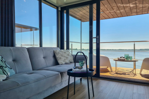 Woonkamer in woonboot met bank en tafel, uitkijkend op terras aan het water in Denemarken.