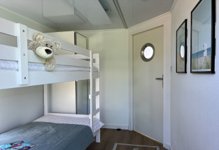 Lichte woonbootslaapkamer met witte stapelbedden, een knuffelbeer en moderne deur met patrijspoort.