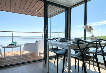 Moderne woonboot in Denemarken met eetruimte en terras met zicht op het water onder een heldere hemel.