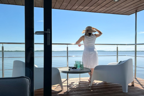 Vrouw in witte jurk met hoed op terras van woonboot voor twee personen genietend van uitzicht in Denemarken.