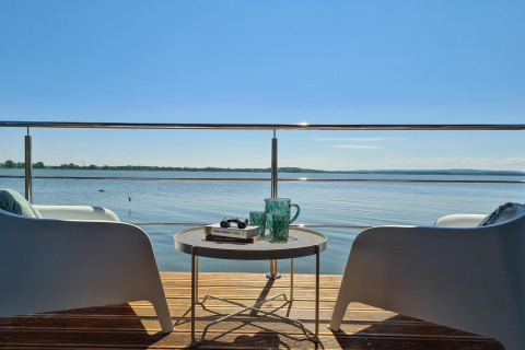 Uitzicht vanaf een woonboot in Denemarken met twee stoelen, tafel, boeken en glazen aan het water.