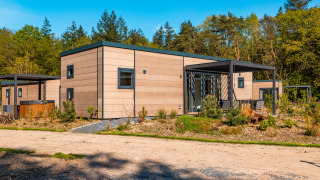 Moderne Groeneborg lodge met hottub, omgeven door bomen en natuur, onder een heldere blauwe hemel.
