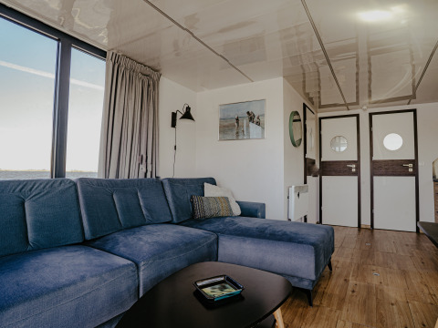 Stue på husbåd 'Campi' ved Marina Uitgeest i Holland med blå sofa og store vinduer mod vandet.