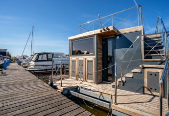 Een moderne woonboot genaamd Houseboat Campi aangemeerd aan een houten steiger onder blauwe lucht.
