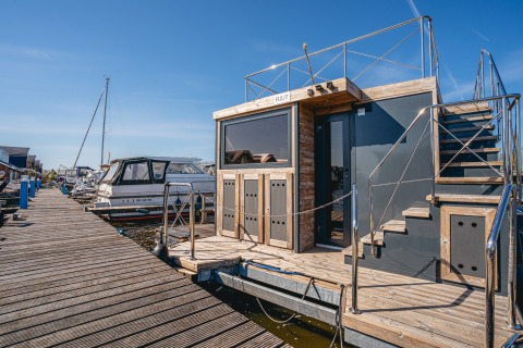Une péniche moderne nommée Houseboat Campi, amarrée à un quai en bois sous un ciel ensoleillé.