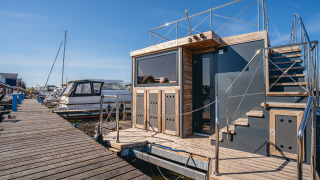 A modern houseboat named Houseboat Campi docked at a wooden pier, with blue sky and nearby boats.