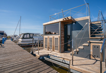 Ein modernes Hausboot namens Houseboat Campi, vertäut an einem Holzsteg an einem sonnigen Tag.