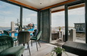 Intérieur moderne du Houseboat Havenlodge avec murs en verre, vue sur l’eau et terrasse ensoleillée.