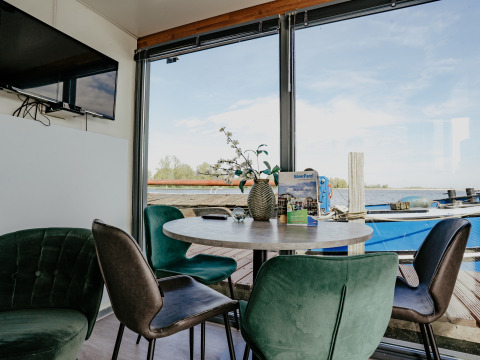 Wohnbereich mit rundem Tisch, grünen Stühlen und Seeblick auf dem Hausboot Havenlodge in Marina Uitgeest.
