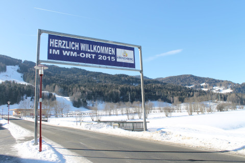 Willkommensschild am Straßenrand in Kreischberg, WM-Ort 2015, mit verschneiten Bergen im Hintergrund.