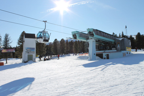Verschneite Skihütte mit Gondelbahn und Sonnenschein, umgeben von Bäumen und blauem Himmel im Winter.