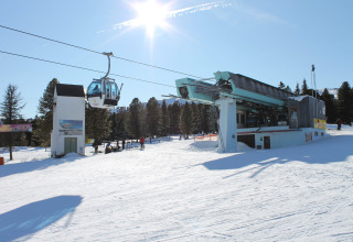 Verschneite Skihütte mit Gondelbahn und Sonnenschein, umgeben von Bäumen und blauem Himmel im Winter.