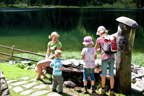 Een familie ontspant bij een berghut aan een meer, terwijl de kinderen buiten in de zon spelen.
