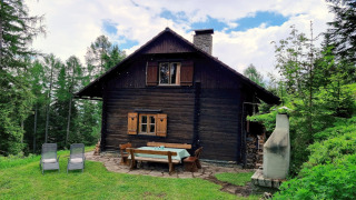 Schneiderhütte-Chalet im Wald mit Holzmöbeln im Garten, Liegestühlen und Grill unter blauem Himmel.