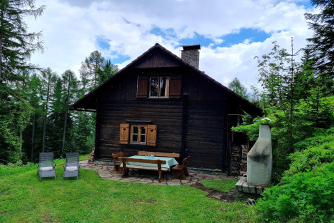 Schneiderhütte-Chalet im Wald mit Holzmöbeln im Garten, Liegestühlen und Grill unter blauem Himmel.