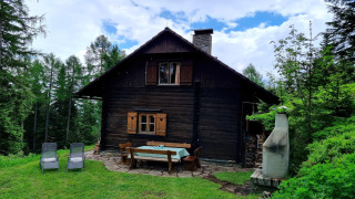 Schneiderhütte chalet in een bos met houten tuinmeubels, ligstoelen en een buitenhaard bij blauwe lucht.