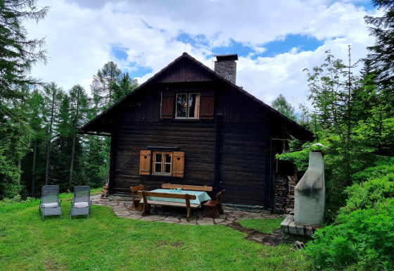 Schneiderhütte-Chalet im Wald mit Holzmöbeln im Garten, Liegestühlen und Grill unter blauem Himmel.